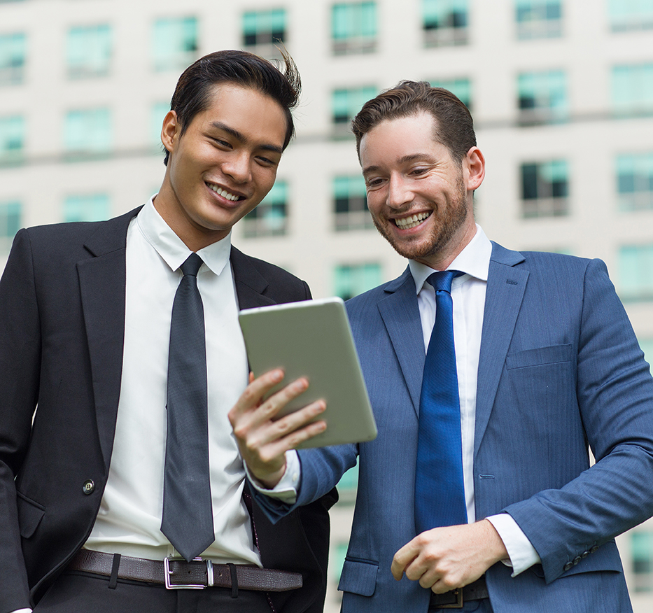 Closeup of two smiling young business men using tablet computer and standing with office building in background